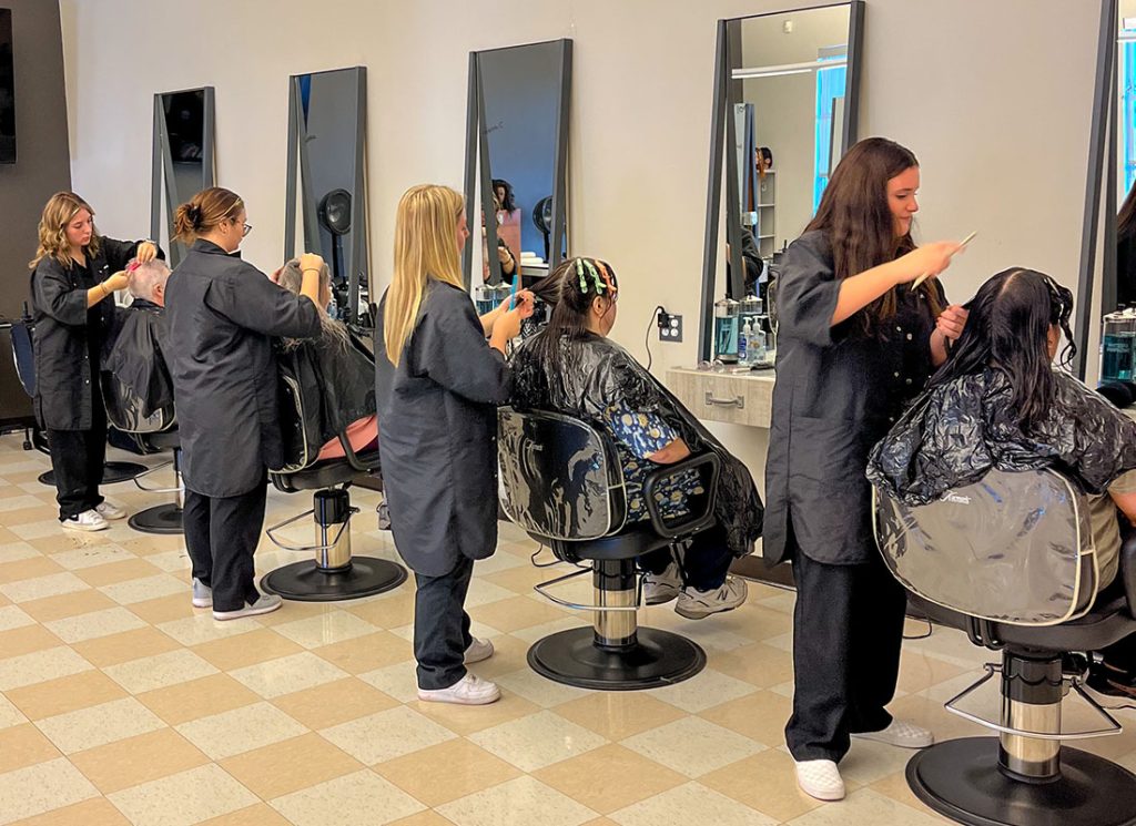 Cuatro estudiantes trabajan en el corte de cabello en cuatro estaciones de trabajo.
