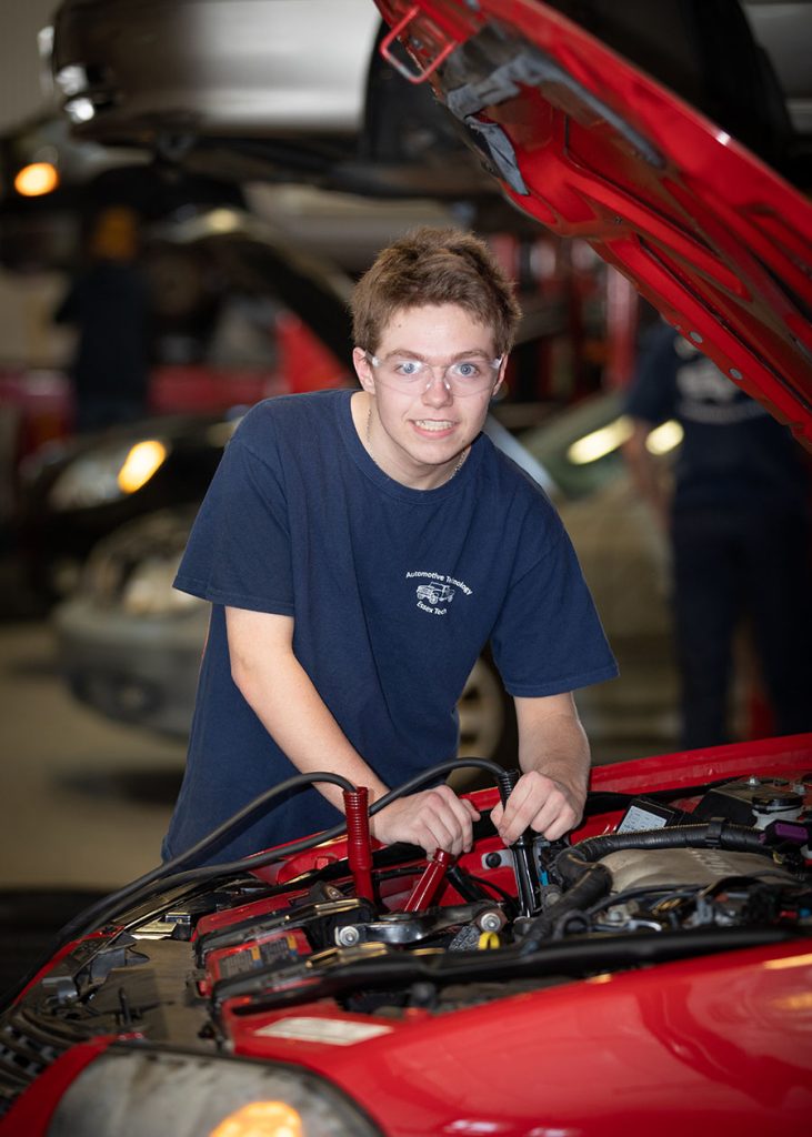 Een student kijkt omhoog naar de camera vanuit de motor van een auto waaraan hij werkt.