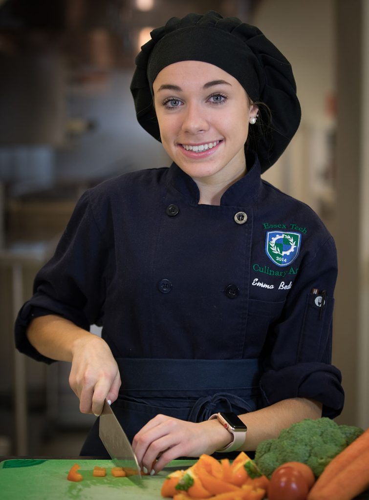 Um aluno com uniforme completo de chef corta vegetais.