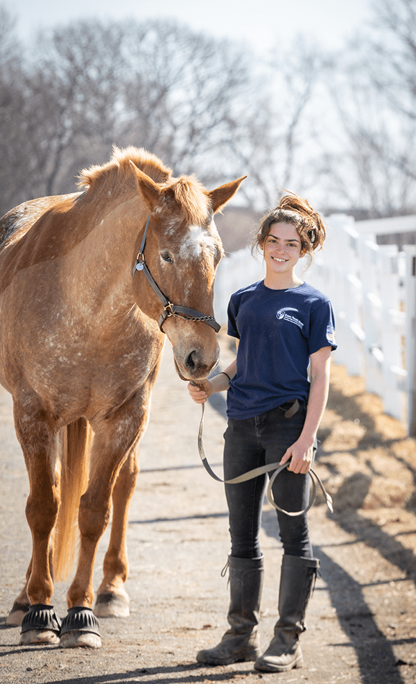 一名学生站在一匹马旁边，牵着马的牵绳，两人都笑眯眯地看着镜头。
