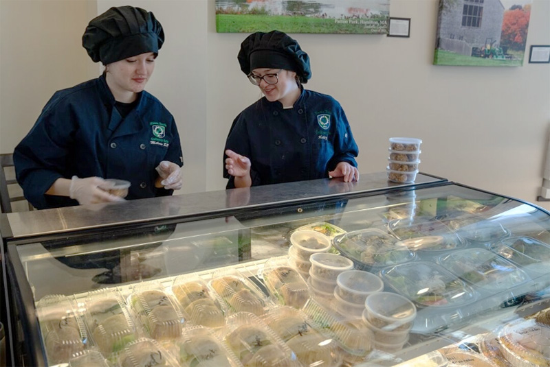 Two students serve sandwiches from behind a glass counter.