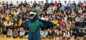 The school mascot - a large blue hawk - stand with wings outstretched in front of students in the gym stands.